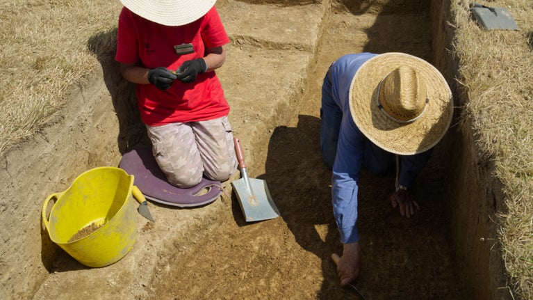 Two people wearing wide-brimmed hats are kneeling in an excavation trench with tools.